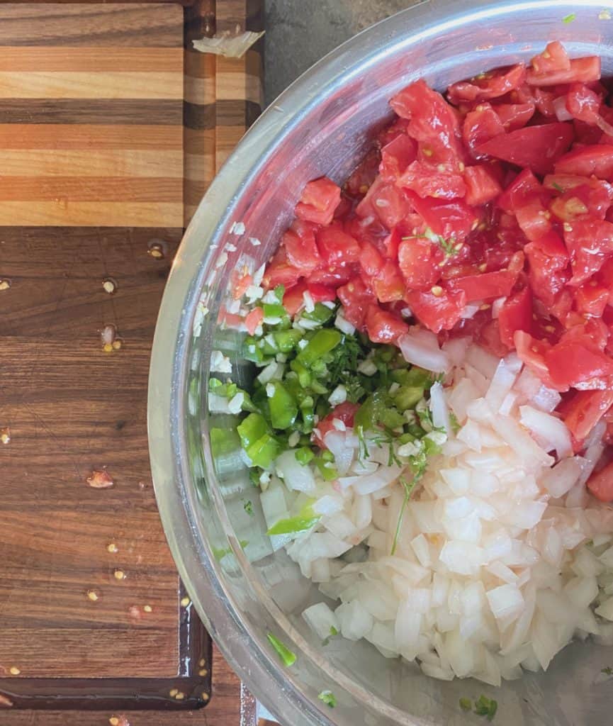 chopped ingredients for pico de gallo in metal bowl with handcrafted carving board beside it