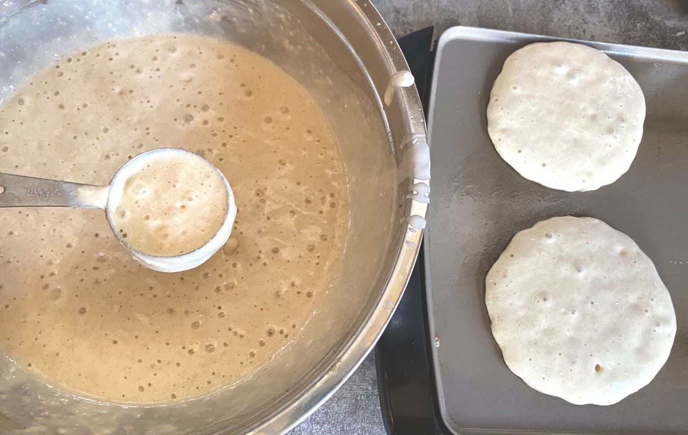 pancake batter in measuring cup with pancakes cooking on griddle beside